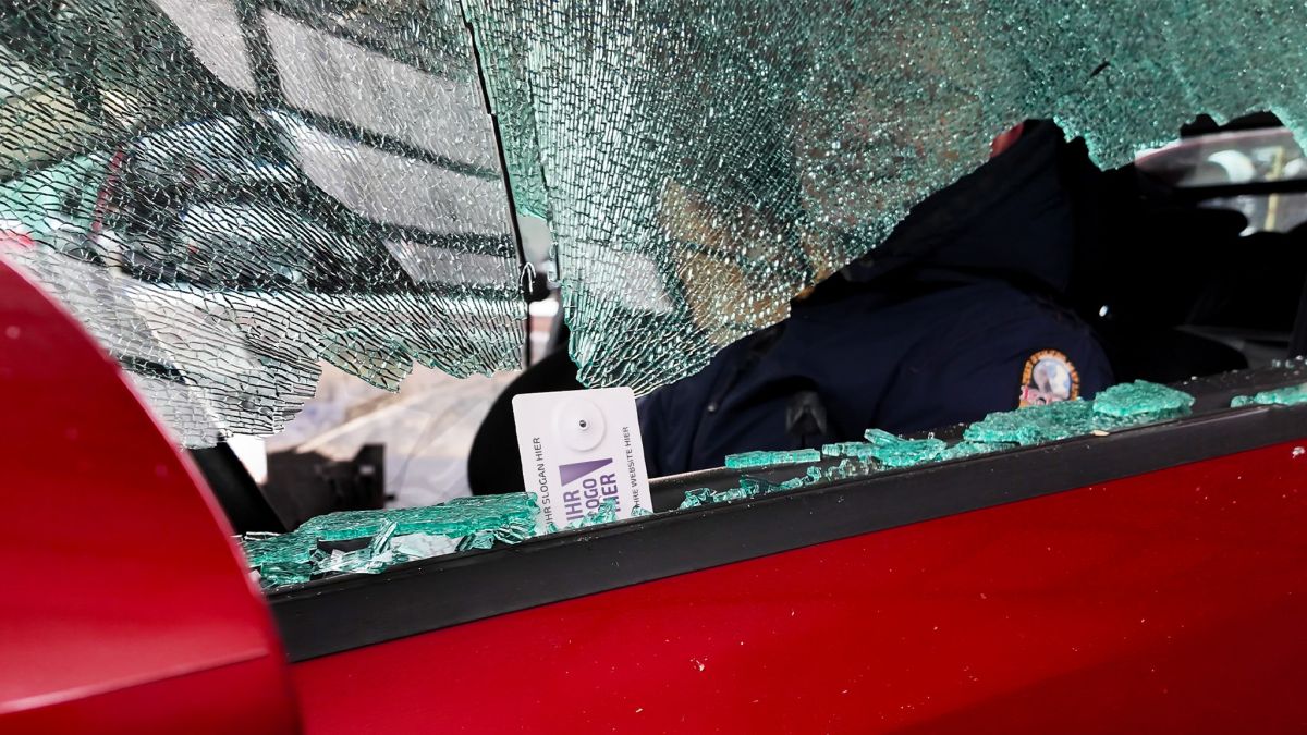 Zerbrochene Autoscheibe mit zersplitterter Autoscheibe und Glasscherben auf roter Tür, Blick ins Innenraum mit Person in dunkler Jacke.