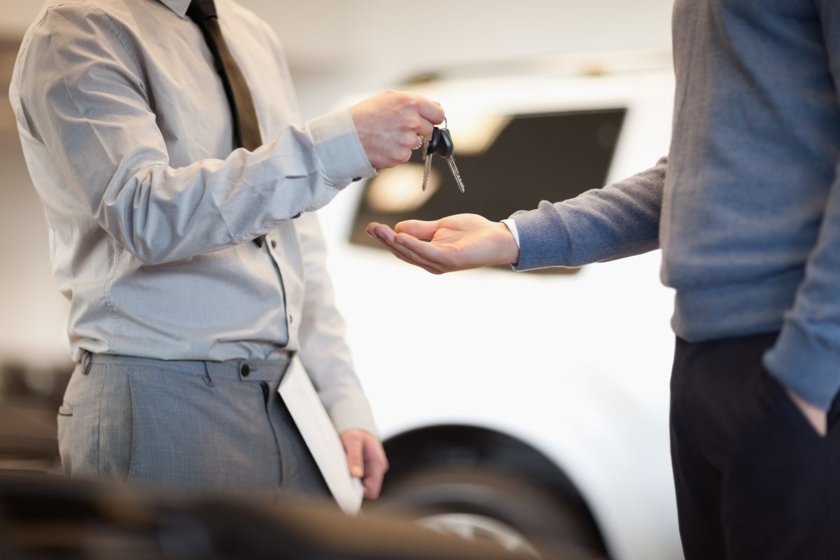 Verkäufer übergibt Autoschlüssel an Käufer, beide in Geschäftskleidung vor einem weißen Auto im Verkaufsraum.