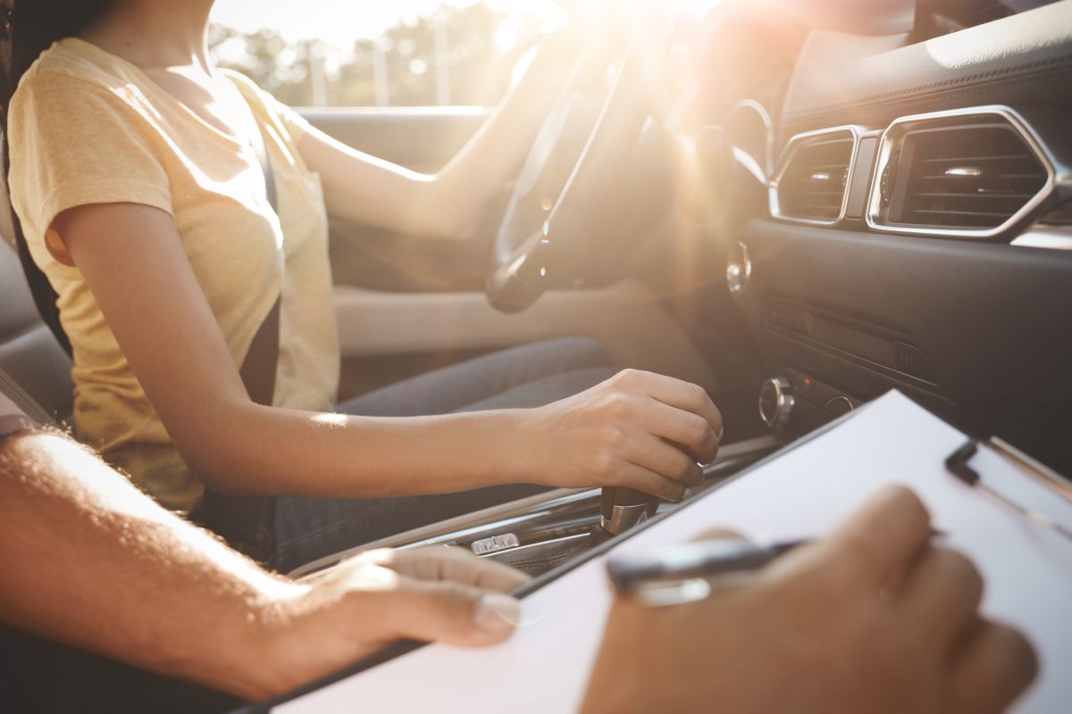 Frau am Steuer eines Autos schaltet mit der rechten Hand, während eine andere Person auf einem Klemmbrett Notizen macht.