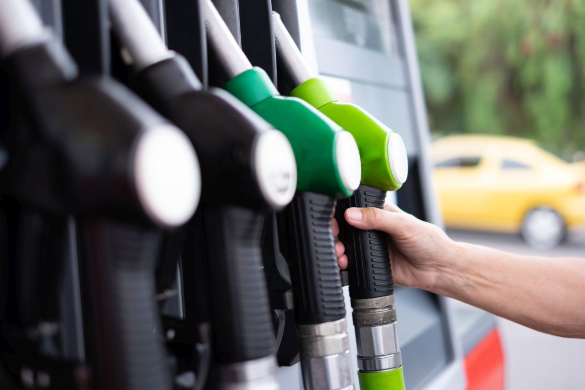 Hand greift nach grünem Zapfventil an einer Reihe von Zapfpistolen an einer Tankstelle, gelbes Auto unscharf im Hintergrund