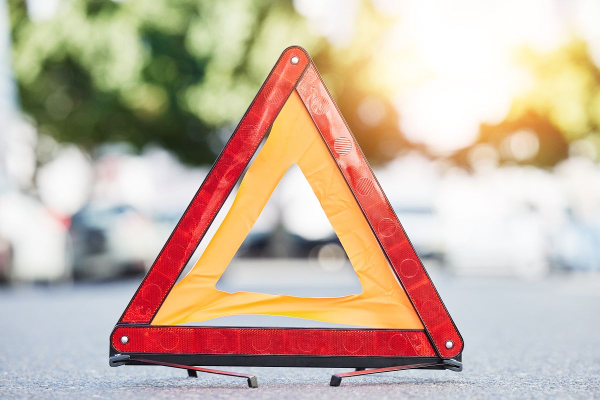 Rotes reflektierendes Warndreieck mit gelbem Innenkeil auf der Straße, unscharfer Hintergrund mit Fahrzeugen und Bäumen