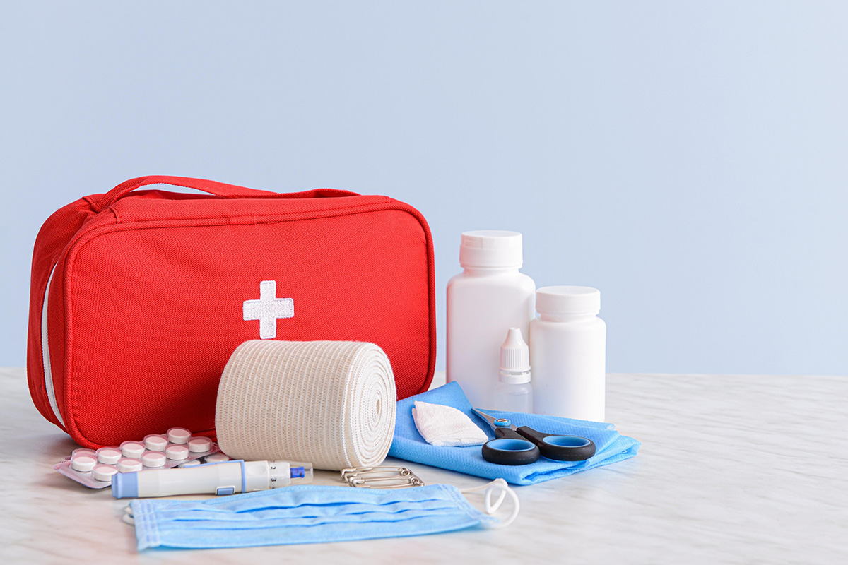 Rotes Verbandskästchen mit weißem Kreuz neben Mullbinde, Pflastern, Tablettenblistern, Masken, Schere, Medikamentenfläschchen und Einmalhandschuhen.
