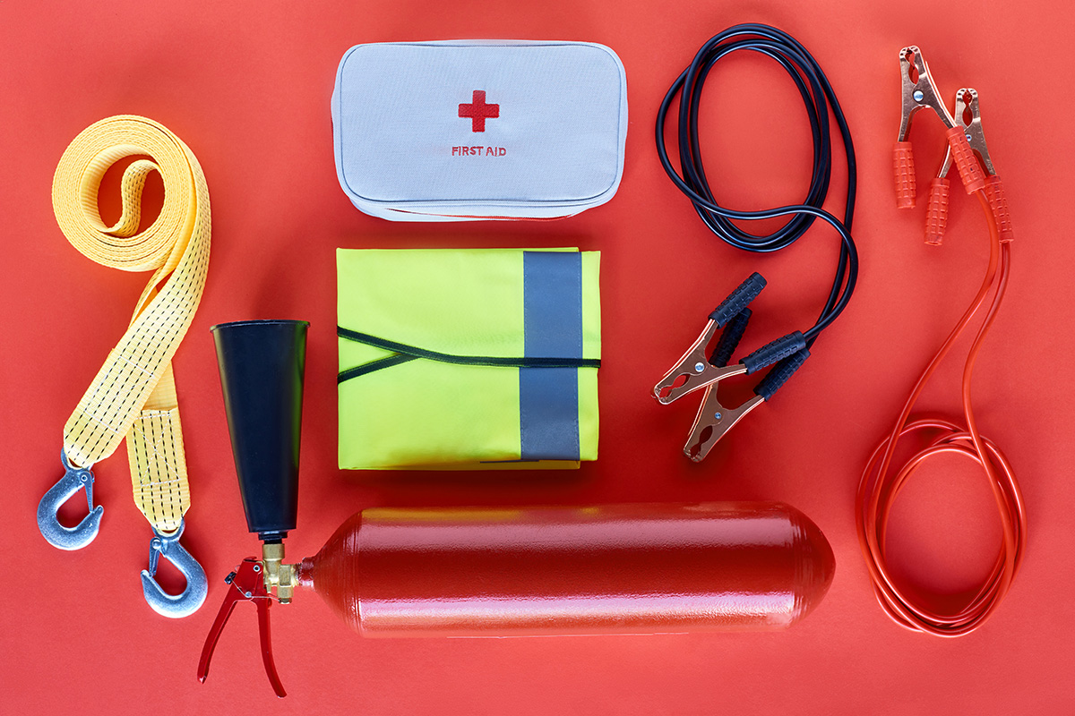 Notfallausrüstung auf rotem Hintergrund: Abschleppschlaufen, Erste-Hilfe-Tasche, Warnweste, Starthilfekabel und Feuerlöscher angeordnet.