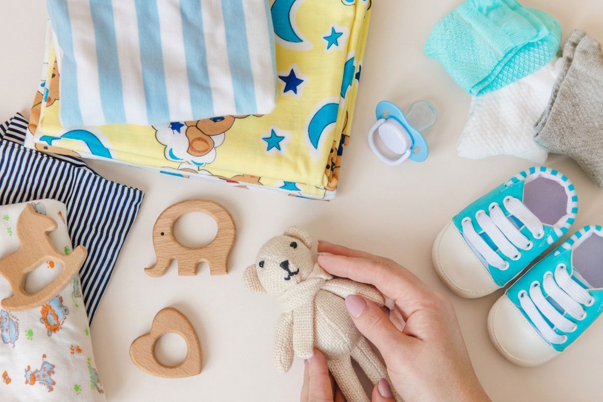Babyartikel auf hellem Hintergrund: Stoffwindeln, Schnuller, türkis-weiße Babyschuhe, Holzspielzeug und Hände halten einen kleinen gestrickten Teddybär.
